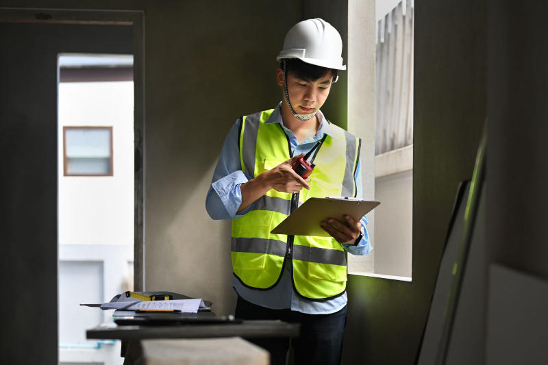 An engineer in a hard hat and safety vest holding a clipboard and checking the progress of a building construction site.