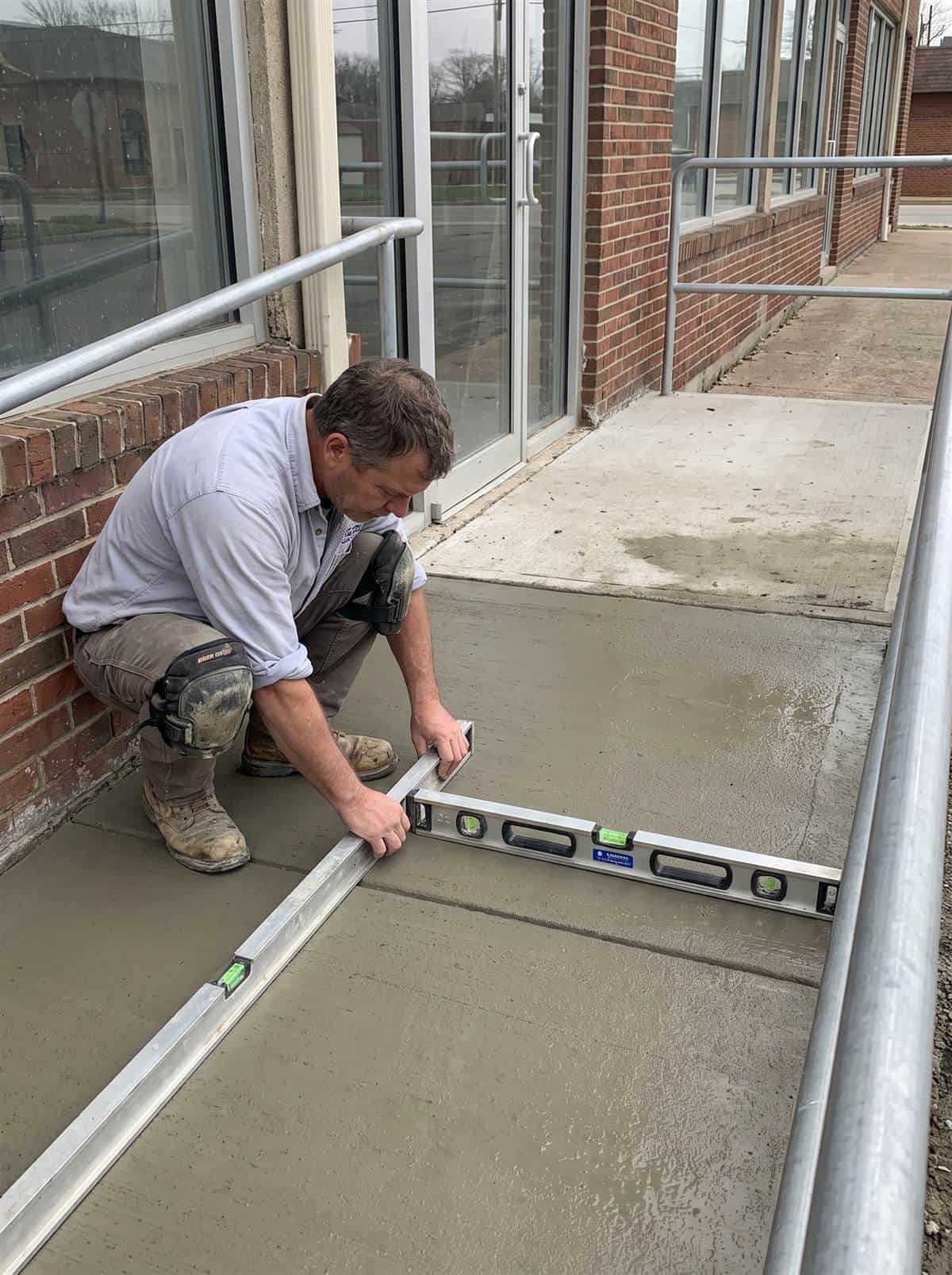 Contractor crouching to check the running slope of a newly finished concrete access ramp with a digital level