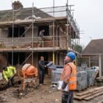 Supervisor with a clipboard overseeing three trades on a scaffolded house extension, a typical CDM site