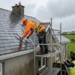 Roofer in a harness replacing slates on a Wexford semi-detached house while a helper steadies the ladder