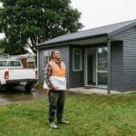 Builder in a hi-vis vest holding rolled plans looks up at a newly finished single-storey weatherboard dwelling on a back section