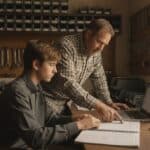 Contractor leans over his teenage son at a shop office desk as the son enters timesheets on a laptop