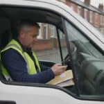 Electrician in a hi-vis vest reads an email on his phone in his van on a terraced street