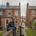 Builder talks over the garden wall with a neighbour behind terraced houses with scaffolding