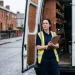 Plumber standing at the open rear doors of her van on a Limerick street, clipboard in hand