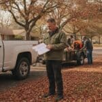 Landscaping company owner reviews figures on a clipboard beside a work truck while two crew members load a leaf blower onto a trailer