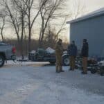 Landscaping crew in work jackets and toques talks beside mowers and a pickup with trailer in a snow-dusted equipment yard