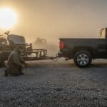 Landscaper kneels to attach trailer safety chains to a pickup in a misty equipment yard