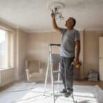 Electrician in a grey t-shirt beside a stepladder pointing at the wiring of an old ceiling rose on a textured ceiling, drill lowered in his other hand, dust sheet on the floor