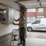 Electrician in a grey work polo lifting a white wall-mounted battery unit into place beside a consumer unit in a semi-detached house garage, a drill on a step ladder and a white van on the drive