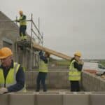 Building crew in hi-vis vests and hard hats laying concrete blocks and carrying timber on a half-built house, scaffolding, a white van and green fields under a grey sky