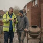 Site manager in a hi-vis jacket checks hours on his phone with a young labourer beside a cement mixer