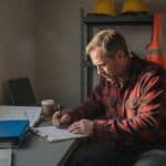 Construction company owner in a red plaid flannel shirt writing on a form on a clipboard at a desk in a site trailer, an open binder and a coffee cup beside him, hard hats and a hi-vis vest on a shelf behind