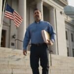 Contractor in a blue work shirt carries a folder of papers down the steps of a county courthouse